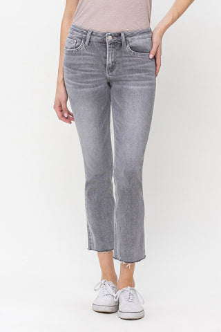 A person wearing VERVET by Flying Monkey's Graylee Mid-Rise Slim Straight Crop Pant in light grey Comfort Stretch Denim with frayed hems, a light pink shirt tucked in, and white sneakers. Their right hand rests on their hip. The background is plain white.