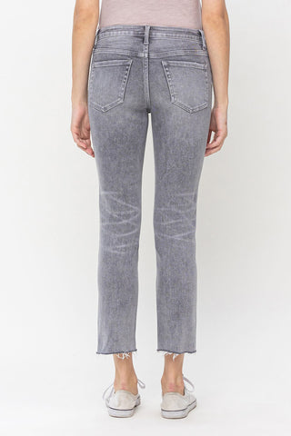 The image shows a person from the back, wearing a light pink top and VERVET by Flying Monkey's Graylee Mid-Rise Slim Straight Crop Pant made of comfort stretch denim with raw, frayed hems. They are also wearing light-colored slip-on shoes.