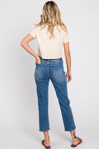 A person with long, wavy blonde hair is standing with their back to the camera. They are wearing a beige short-sleeve top, Petra153's Mavee High Rise Stretch Slim Crop Jeans in blue, and brown sandals. They have one hand resting on their hip, and there is a small tattoo visible on the back of their left ankle.