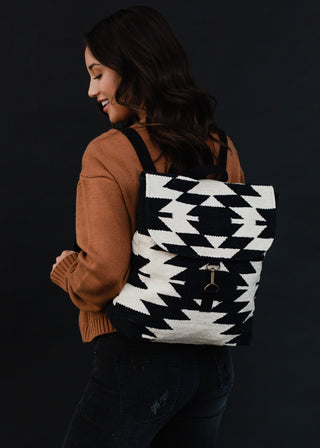 A woman with long dark hair wears a brown sweater and black jeans. She is smiling and looking down over her shoulder, showcasing the Quaid Woven Backpack from Panache Accessories, featuring intricate black and white Aztec geometric patterns. The dark background serves to highlight both her and the striking backpack.