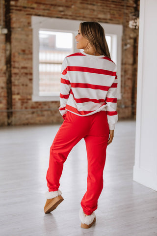 A person stands indoors against light floors and exposed brick walls, wearing a Callahan Striped Pullover and Joggers Set by Liam & Company. The ensemble features a red and white striped top, matching joggers, and brown slippers with white fluffy trim.