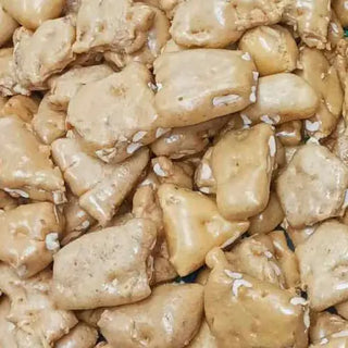 A close-up of a pile of Western Edge Boutique's Homemade Peanut Brittle 4oz showcases its distinctive texture, featuring a glossy caramel coating with visible peanut pieces embedded throughout. The brittle pieces vary in size and shape, creating a textured surface reminiscent of freeze-dried candies.