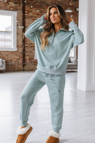 A woman stands inside a stylish room with exposed brick walls. She wears a relaxed fit, light blue Alison Half Zip Loungewear Set from Liam & Company, paired with cozy brown slippers lined with white fur. She has long, wavy hair and is cheerfully posing with her hands near her face.