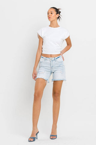 A person stands against a white background, wearing a white T-shirt, Kolson Mid Rise Distressed Stretch Denim Shorts in light blue from VERVET by FLYING MONKEY, and blue high-heeled sandals. They have their hair tied back and one hand resting on their hip.