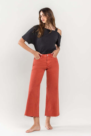 A woman with long, wavy hair is wearing an off-shoulder black top and the VERVET by Flying Monkey Scout High Rise Crop Wide Leg Jeans in a rust color. She stands barefoot against a plain, light-colored background, with one hand in her pocket and a relaxed expression.