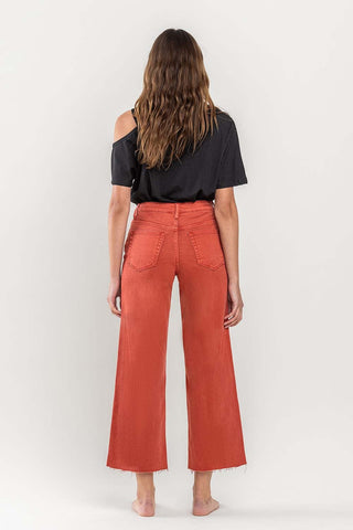 A person with long hair stands barefoot, facing away from the camera. They wear an off-the-shoulder black t-shirt and VERVET by Flying Monkey's Scout High Rise Crop Wide Leg Jeans in orange, featuring frayed hems and made of comfort stretch denim. The background is a plain, off-white color.