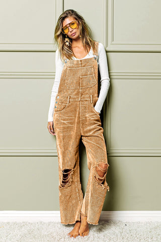 A person leans against a light green paneled wall, wearing a white ribbed long-sleeve top under the BiBi Mav Distressed Corduroy Overalls with ripped knees. They have loose, wavy hair and wear yellow-tinted sunglasses. They stand barefoot on a plush, cream carpet.