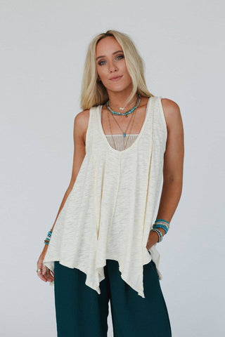 A woman with long blonde hair stands against a plain background, wearing the Elara Top - Neutral from Three Bird Nest, a loose-fitting, boho-style white sleeveless top with a handkerchief hem. She accessorizes with layered turquoise and silver necklaces and bracelets, and pairs the comfy top with dark green pants.