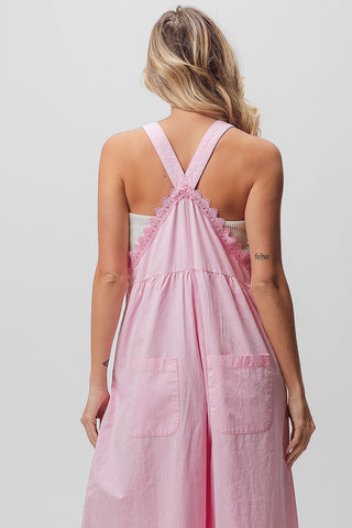 A person with long blonde hair is shown from the back wearing the BiBi Tate Lace Edge Detail Wide Leg Overall, which is a light pink, shoulder-strapped outfit made of lightweight cotton. The overall features lace detailing at the top and two large pockets on the lower back. The person has a small tattoo on their upper left arm.