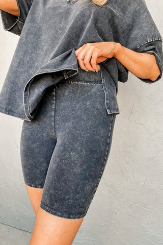 A person wearing the Harper Gray Acid Loungewear Set from Pretty Bash stands against a textured, light gray background. They lift the hem of their oversized shirt slightly, enhancing the retro style of their look.