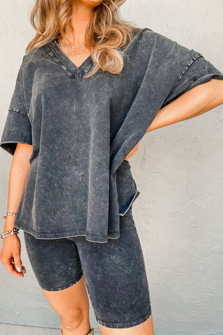 A person with long, wavy hair wears the Harper Gray Acid Loungewear Set from Pretty Bash, featuring an oversized acid-wash T-shirt with a V-neck and matching biker shorts, embracing a retro style. They accessorize with beaded bracelets on one wrist and a layered necklace. Their pose has one hand resting on their hip.