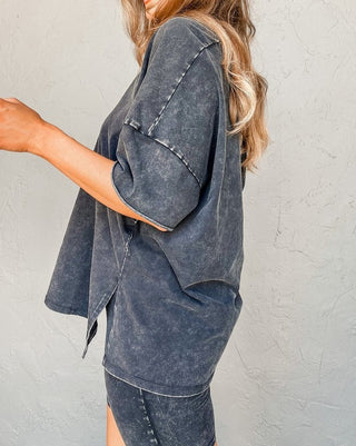 A person with long, wavy hair stands against a light textured wall, seen from the side. They wear an oversized Harper Gray Acid Loungewear Set from Pretty Bash with rolled-up sleeves. Their arm extends forward, partially out of the frame, giving a relaxed vibe to their comfortable cotton/polyester blend outfit.