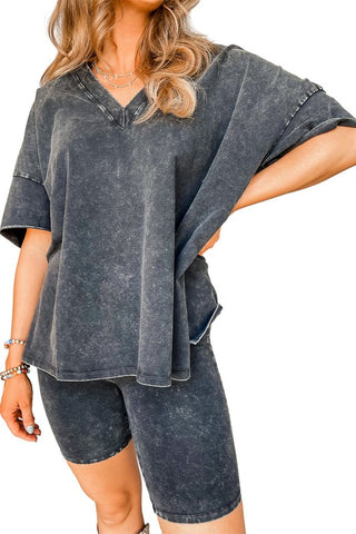 A person is wearing the Harper Gray Acid Loungewear Set by Pretty Bash, featuring a loose, dark acid-washed V-neck T-shirt paired with matching high-waisted biker shorts. They have long, wavy hair and are accessorized with layered necklaces and bracelets. With their arm bent at the elbow and hand resting on their hip, they embody a cool retro style.