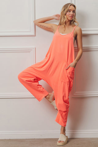 A person poses against a white paneled wall, wearing the loose and vibrant Hattie One Piece Soft Loungewear in bright coral from bibi—an absolute weekend must-have. They have one leg bent, with their foot resting against the other leg, and one hand casually tucked in their pocket. Standing on a wooden floor with light-colored slide sandals, tattoos are visible on their arm.