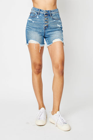 A person wearing the Elsi Rigid Magic Destroy & Button Fly Cut Off Shorts from Judy Blue stands against a plain white background, completing the outfit with white sneakers.