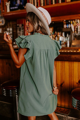 A woman in a Hallie Olive Green Baby Doll Ruffle Sleeve dress by Western Edge Boutique and a white hat stands at a wooden bar, holding a drink. The softly blurred background showcases bottles, creating a warm, rustic atmosphere. Her dress features charming ruffle sleeves, adding an elegant touch to her summery look.