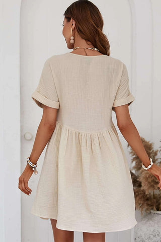 A woman with long brown hair stands facing a white arched doorway, wearing the stylish Jordy Short Sleeve Beige Lace V Neck Dress from Western Edge Boutique. She is accessorized with bracelets and a necklace, and has large earrings. Dried plants are visible to her right.