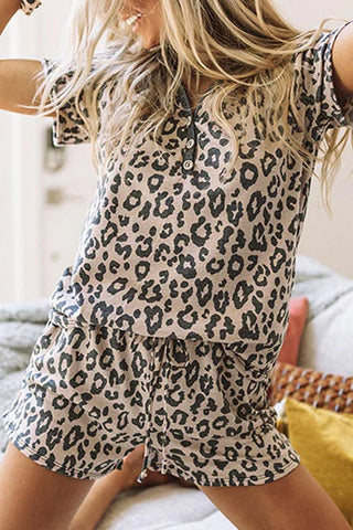 A person dressed in the stylish Laddi Leopard Lounge Set from Western Edge Boutique stands with arms raised in excitement or joy. The scene is set indoors, where a couch is visible in the background, perfectly merging fashion with comfort.