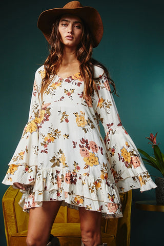 A woman with long wavy hair wearing a floppy brown hat and a Western Edge Boutique Lacey White Printed Floral Print Layered Sleeve Dress stands against a dark green background. The dress, adorned with vibrant patterns, features flared long sleeves and a ruffled hemline at the bottom.