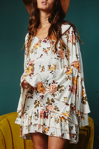 A person wearing a Lacey White Printed Floral Print Layered Sleeve Dress from Western Edge Boutique. The individual has long, wavy hair and is accessorizing with a wide-brimmed hat. The background includes a green wall and a mustard-yellow chair.