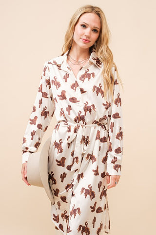 A woman with long blonde hair exudes style in the Satin Western Print Button Up Shirt Dress by Western Edge Boutique, showcasing a cream-colored design adorned with a brown pattern of cacti and cowboy hats. She holds a light-colored hat in her right hand against a beige background.