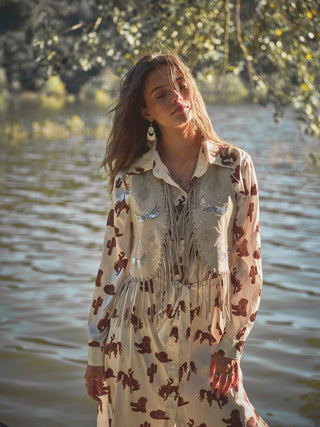 A woman stands in front of a lake, wearing the Satin Western Print Button Up Shirt Dress from Western Edge Boutique, along with a fringed faux leather vest. Sunlight filters through the trees as her eyes are closed, and her hair gently blows in the breeze.