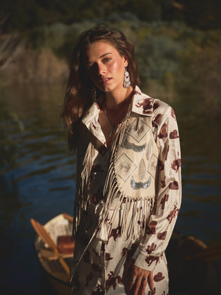 A person stands near a body of water dressed in a patterned outfit and accented by the Faux Leather Embellished Fringe Vest from Western Edge Boutique, complete with large earrings. The western-style ensemble is enhanced by intricate bead artwork on the vest. In the background, greenery and a partially visible boat can be seen under warm lighting with shadows adding depth to the scene.