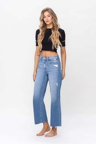 A person with long, wavy hair is wearing a black cropped top and Western Edge Boutique's "Maverick Wide Leg High Rise Ankle Crop Jeans" in light blue with slight distressing. They are standing barefoot against a plain white background.