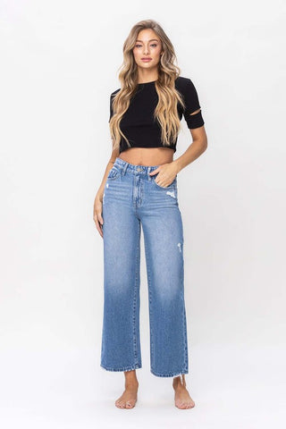 A woman with long, wavy hair stands against a plain background. She is wearing a black crop top and the Maverick Wide Leg High Rise Ankle Crop Jeans from Western Edge Boutique. She is barefoot and has a relaxed, confident expression.