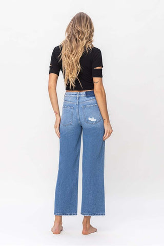 A person with long, wavy blonde hair is standing barefoot, facing away from the camera. They are wearing a black short-sleeved top and the Maverick Wide Leg High Rise Ankle Crop Jeans from Western Edge Boutique. The white background is plain and uncluttered.