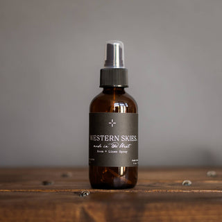 A 4 oz. brown glass spray bottle labeled "Western Skies Room + Linen Spray" by R. Rebellion, infused with sandalwood and vanilla notes, sits on a wooden surface against a gray background.