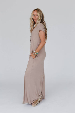 A person with long, wavy blonde hair stands in profile against a plain background, wearing the Kendrick Mocha Jumpsuit from Three Bird Nest. The jumpsuit is light pink, comfy with short sleeves and pockets. They accessorize with several colorful bracelets and wear beige sandals.