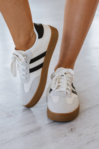 Close-up of a person wearing Ness Striped Lace Up Flat Sneakers: White Black from Liam & Company. The sneakers feature brown soles and are laced up, and the person is standing on a light-colored wooden floor.