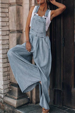 A person leans against a wooden door frame, wearing the Honeypeach Everson Lightweight Denim Wide Leg Denim Overall in light blue over a white sleeveless top. One leg is crossed over the other, and their left arm is bent with the hand resting on their head. The outfit exudes a casual, relaxed style.