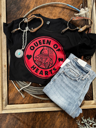 A "Queen Of Hearts" graphic tee from Western Edge Boutique, featuring a red card design, is placed on a wooden surface. Blue jeans, a silver necklace with turquoise pendant, and brown leather accessories are artistically arranged alongside a rope.