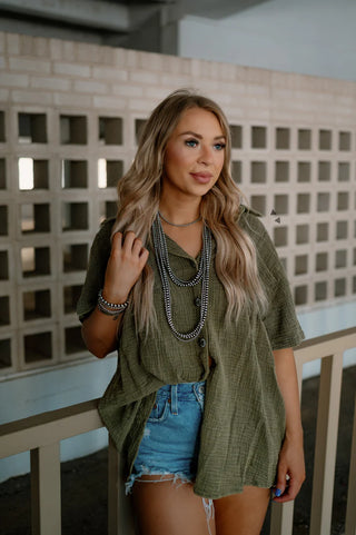 A woman with long blonde hair stands in front of a wall with rectangular cutouts. She is wearing a **RODEO GROUNDS *AGAVE** loose, oversized fit green shortsleeve button-up shirt by **2 Fly** and denim shorts. She accessorizes with layered necklaces and bracelets.