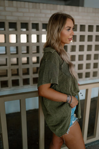 A woman with long blonde hair stands in profile, wearing the 2 Fly RODEO GROUNDS *AGAVE oversized olive green shirt with rolled-up short sleeves. She pairs it with distressed denim shorts and multiple bracelets adorn her wrist. The background features a brick wall with square cutouts.