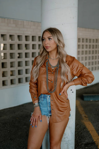 A woman with long blonde hair stands against a pillar. She wears a Western Edge Boutique's Leather Lux Saddle Brown Long Sleeve Top with turquoise pearl snaps and distressed denim shorts. She accessorizes with multiple layered necklaces, bracelets, and has blue nail polish. The background features a beige block wall.