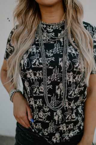 Person with long, wavy hair wearing the Dusk Desert Top by 2 Fly, featuring a white cowboy and cactus print on super soft black fabric. Accessorized with multiple beaded necklaces. Only the torso and part of the lower face are visible.