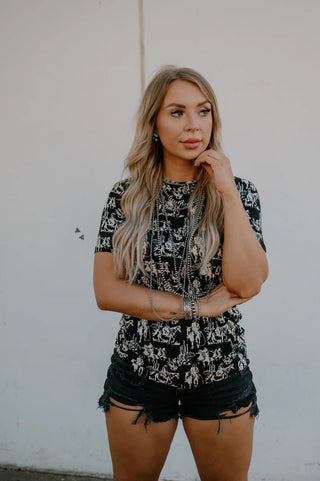 A woman with long blonde hair stands against a plain white wall. She is wearing a black and white patterned Dusk Desert Top from 2 Fly, made from super soft fabric, paired with black denim shorts. She has multiple bracelets on her wrist and one hand is touching her chin.