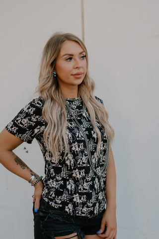 A woman with long, wavy blonde hair stands against a white wall. She is wearing the Dusk Desert Top by 2 Fly, a black and white patterned t-shirt made from fabric with lots of stretch, paired with distressed black denim shorts. She has blue nail polish, various bracelets, and a visible tattoo on her left arm. She gazes off to the side.
