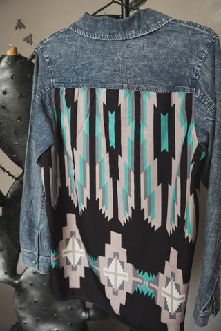 The Grand Prairie Chambray Long Sleeve Shirt by 2 Fly is displayed hanging on a hanger. With a colorful geometric patterned back showcasing black, white, teal, and beige shapes along with turquoise snaps, it exudes Southwestern charm. The indoor setup includes decorative elements like faux cactus plants and metallic insects on the wall.