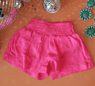 A pair of Maddi Careless Fuchsia Shorts by 2 Fly, featuring an elastic yoga band waistband, is placed on a brown surface. The scene is accentuated with decorative items including disco balls and turquoise and silver jewelry pieces, creating a stylish and colorful ensemble.