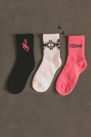 Three SOCK EM SILLY POP socks from the brand 2 Fly are displayed side by side against a dark background. The black sock showcases a pink cowboy rider graphic, the white sock features a black geometric pattern, and the pink sock has "YEEHAW" written in bold black text. Two small flies hover near the white sock. This is a perfect 3-pack crew socks set for any western enthusiast; one size fits most.