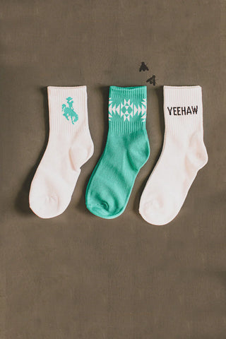 Three pairs of SOCK EM SILLY *CRISP western socks from 2 Fly are displayed on a flat surface. The left pair is white with a turquoise horse graphic, the middle pair is green with a white geometric pattern, and the right pair is white with the word "YEEHAW" printed in black text. This 3 pack comes in one size that fits most.