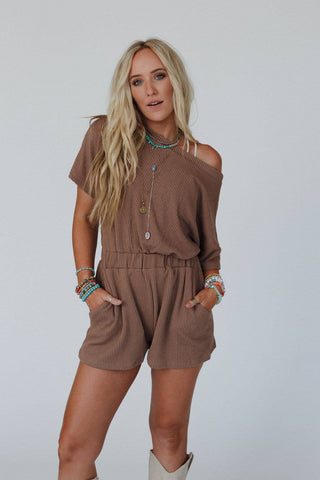 A woman with long, wavy blonde hair poses confidently with her hands in the pockets of her camel-colored, off-the-shoulder Covey Romper from Three Bird Nest. Featuring an elastic waistband and ribbed knit fabric, she accessorizes with layered necklaces, bracelets, and beige cowboy boots against a plain white wall backdrop.