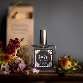 A bottle of R. Rebellion's Coastal Cowgirl Perfume 50 ml sits on a table next to dried flowers. The bottle, featuring a silver cap and adorned with a decorative label showcasing botanical designs, promises a unique and playful scent. Behind the West Coast fragrance, there is a minimalist cardboard box partially blurred in the background.