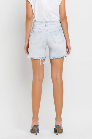 A person stands with their back facing the camera, sporting a white t-shirt and Kolson Mid Rise Distressed Stretch Denim Shorts by VERVET by FLYING MONKEY, complete with light blue frayed denim. Blue high-heeled shoes add a modern touch to this retro look, all set against a plain white background.