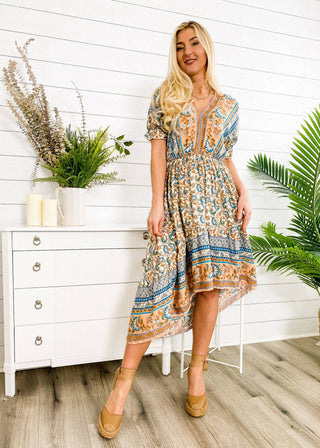 A woman in a Finn Tie Back High Low Dress by Anna Kaci Wholesale stands indoors, smiling. She wears tan wedge shoes and has long blonde hair. Behind her is a white dresser adorned with green potted plants, candles, and decorative items. The wooden floor and white paneled wall complete the setting.