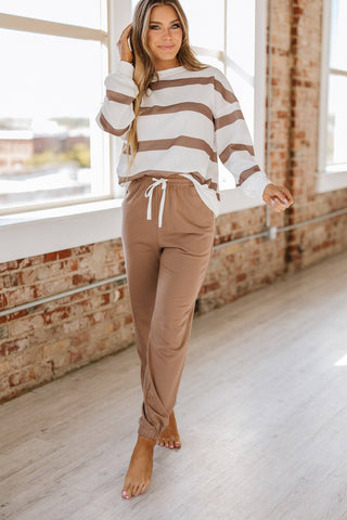 A woman stands indoors on a light wooden floor, wearing the Callahan Striped Pullover and Joggers Set by Liam & Company. Brick walls and large windows fill the space with natural light.